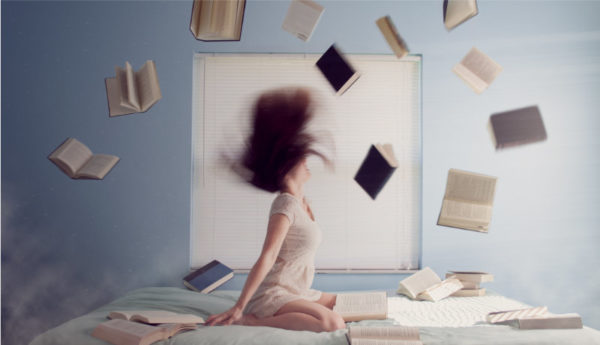 woman sitting on bed head tossed back with hair in air and lots of books thrown up in the air around her