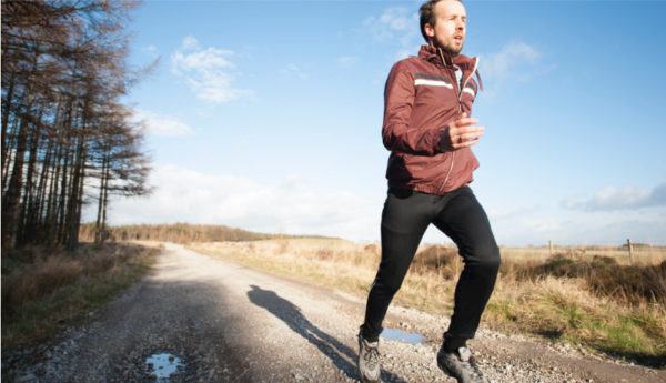man running in sunshine with woods on his right and fields on his left, wearing maroon tracksuit top and black leggings