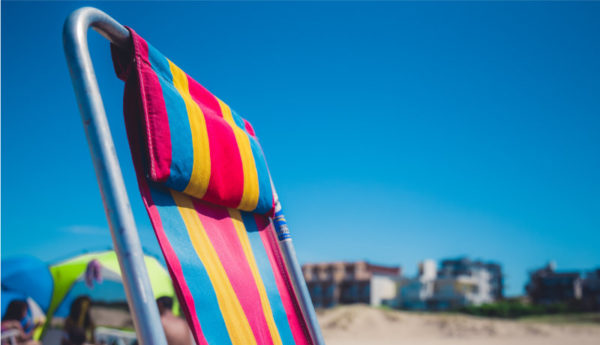 Top half of a deck chair with bright pink, blue and yellow stripes against a clear blue sky and some sand and buildings in the background but hazy