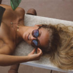 top half of a tanned woman wiht blond hair and black bikini, lying down looking to the side touching her sunglasses